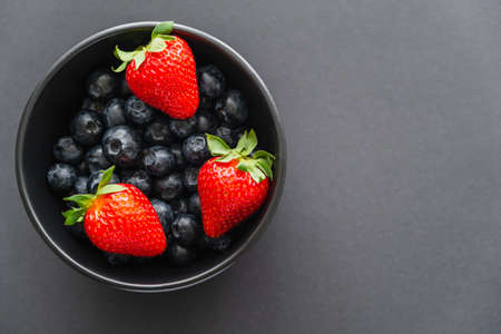 Top view of juicy strawberries on blueberries in bowl on black background
