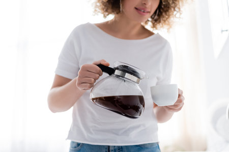 cropped view of curly woman in white t-shirt pouring morning coffee