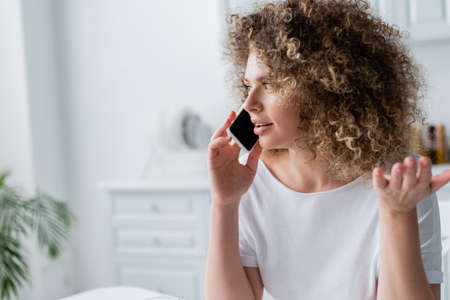 curly woman in white t-shirt gesturing while talking on smartphone at home