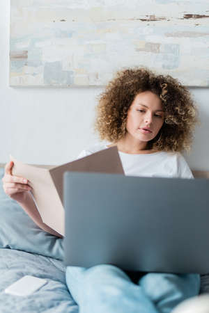 woman with wavy hair holding folder near laptop in bedroomの写真素材