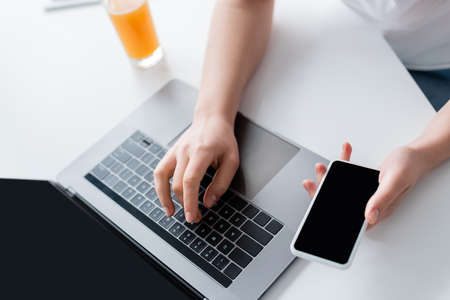 partial view of woman with mobile phone typing on laptop near glass of orange juice
