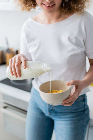 partial view of smiling woman pouring milk into bowl with tasty corn flakes