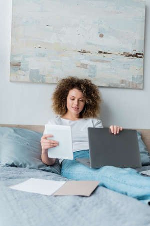 smiling woman with laptop and digital tablet sitting on bed near documents