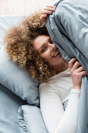 cheerful curly woman looking at camera while hiding under grey blanketの写真素材