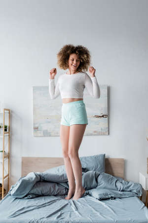 full length of excited curly woman in white pajamas standing on bed at homeの写真素材