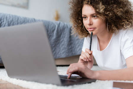 smiling woman holding pen and using laptop on floor in bedroom