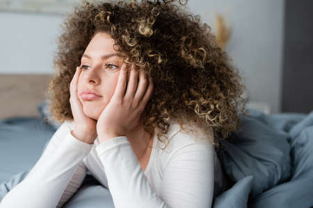 sad and thoughtful woman with hands near face looking away in bedroomの写真素材
