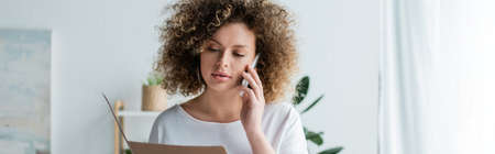 young and curly woman with folder talking on mobile phone at home, banner
