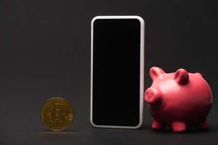 KYIV, UKRAINE - APRIL 26, 2022: Close up view of smartphone near piggy bank and bitcoin on black backgroundのeditorial素材