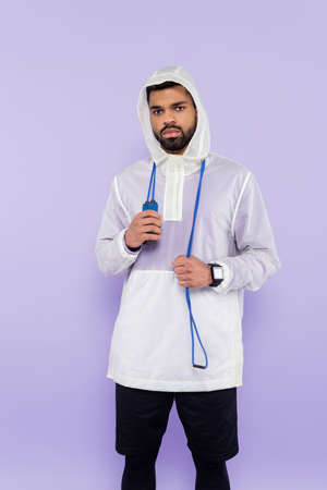 african american sportsman in sportswear with hood looking at camera and holding jumping rope isolated on purpleの写真素材