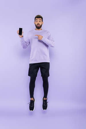 full length of surprised african american man pointing at smartphone and levitating on purpleの写真素材