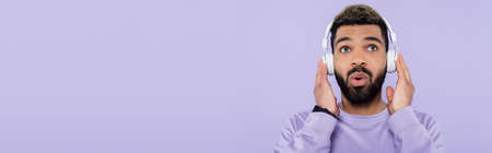 surprised and bearded african american man in wireless headphones isolated on purple, bannerの写真素材