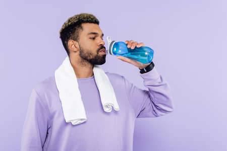 young african american man in sweater standing with white towel and drinking water isolated on purpleの写真素材