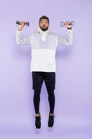 full length of shocked african american man in sportswear levitating and holding dumbbells on purpleの写真素材