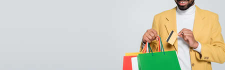 cropped view of cheerful african american man in yellow blazer holding shopping bags and credit card...