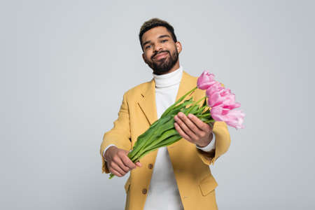 pleased african american man in stylish outfit holding bouquet of pink tulips isolated on greyの写真素材