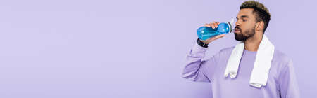 bearded african american man in sweater standing with white towel and drinking water isolated on purple, bannerの写真素材