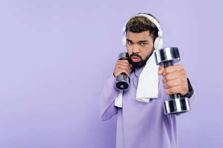 sportive african american man in headphones exercising with dumbbells isolated on purpleの写真素材