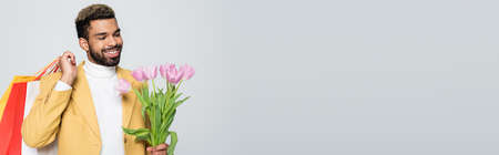 happy african american man in yellow blazer and polo neck holding pink tulips and shopping bags isolated on grey, bannerの写真素材