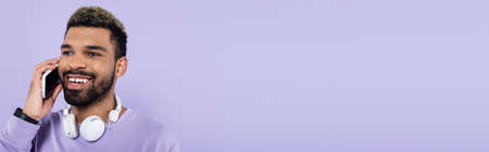 happy african american man in wireless headphones talking on smartphone isolated on purple, banner