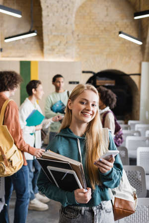 happy woman with notebooks, digital tablet and smartphone near blurred multiethnic friends in auditoriumの写真素材