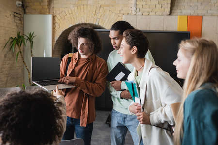 curly man holding laptop with blank screen near friends with notebooks in university