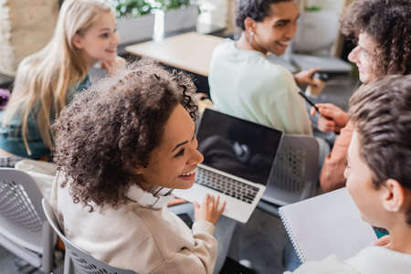 cheerful african american student with laptop looking to blurred friend in classroomの写真素材