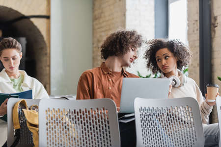thoughtful african american woman with coffee to go looking at curly friend with laptop in...