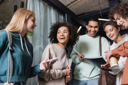 smiling african american man showing copybook to excited interracial students