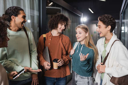 cheerful multicultural students talking and gesturing in university hallway