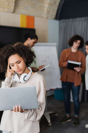 pensive african american woman with laptop and headphones near blurred interracial studentsの写真素材