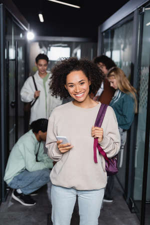 young african american woman with smartphone and backpack smiling at camera near blurred friendsの写真素材
