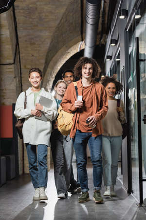 full length of smiling interracial students with gadgets walking along hallway in university