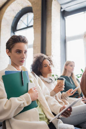 young woman with copybooks and gadgets listening lecture near interracial classmatesの写真素材