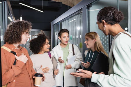 astonished multicultural students gesturing during conversation in university hallの写真素材