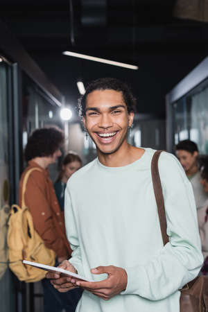 happy and stylish african american student holding digital tablet near people on blurred backgroundの写真素材