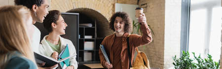 curly man toasting with paper cup near cheerful interracial students, bannerの写真素材
