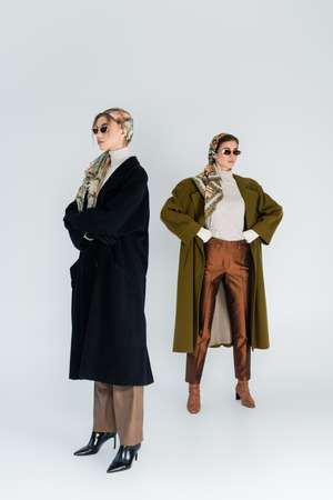 full length of women in coats and sunglasses posing with crossed arms and hands on hips on grey...