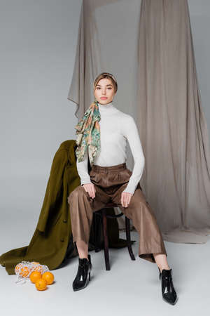 elegant woman sitting on chair near green coat and fresh oranges on grey background with drapery