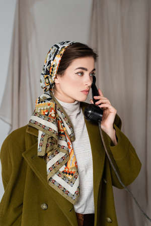 stylish woman in patterned headscarf calling on vintage telephone on grey background with drapery