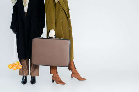 partial view of women in coats holding vintage suitcase and net bag with oranges on grey background