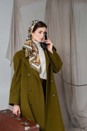 woman in green coat and patterned kerchief holding suitcase and talking on vintage phone on grey...