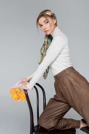 stylish woman in turtleneck and kerchief posing with chair and net bag with oranges isolated on greyの写真素材