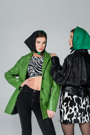 trendy woman in green leather jacket and animal print top posing near friend isolated on greyの写真素材
