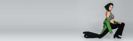 full length of woman in kerchief and black jeans posing on one knee on grey background, bannerの写真素材