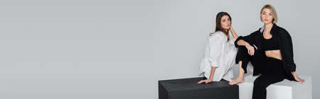 barefoot women in trendy clothing sitting on black and white cubes isolated on grey, banner