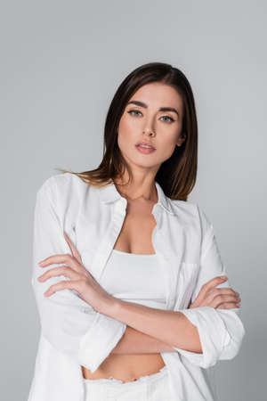 brunette woman in white shirt crossing arms and looking at camera isolated on greyの写真素材