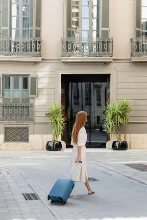 full length of young redhead woman in dress walking with luggage on european street in Valenciaの写真素材