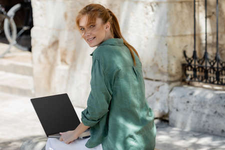 cheerful freelancer sitting on concrete bench and using laptop with blank screen