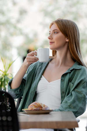 redhead woman holding cup of coffee near tasty croissant on summer terrace